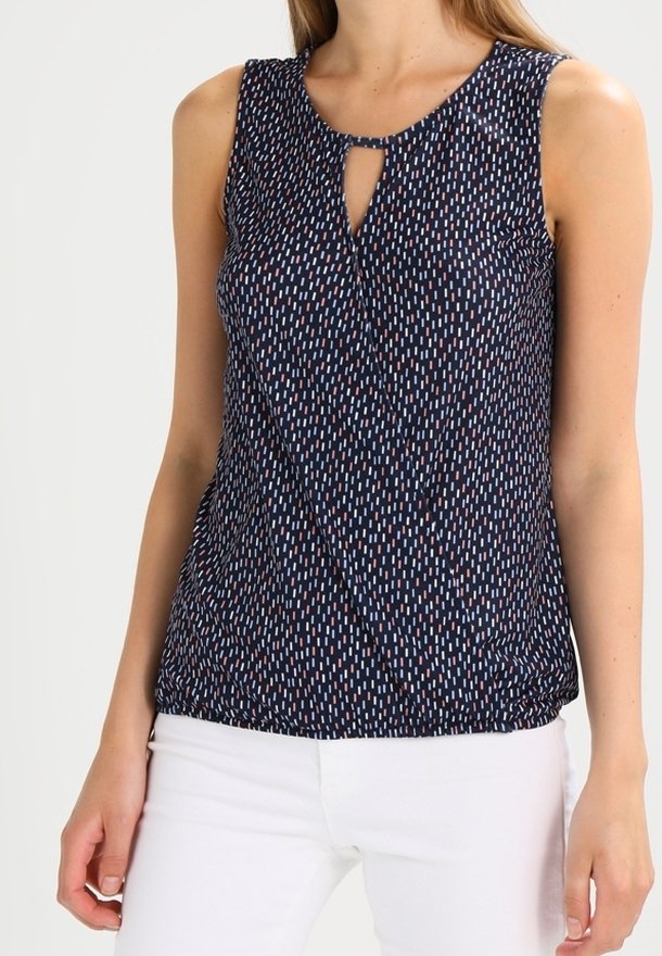 Femme portant un chemisier sans manches bleu marine avec des motifs verticaux en traits blancs et roses, et un pantalon blanc, debout devant un fond uni.