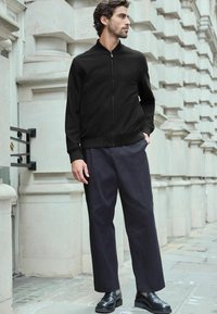 Juodas zip-up švarkas su imitaciniu apykakle, derinamas su laisvo kirpimo tamsiomis kelnėmis ir juodais odiniais batais, prieš tekstūruotą akmeninį foną.