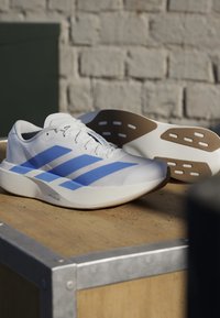 Scarpe da corsa bianche con strisce blu e dettagli marroni sulla suola, appoggiate su una superficie in legno con sfondo di muro di mattoni.