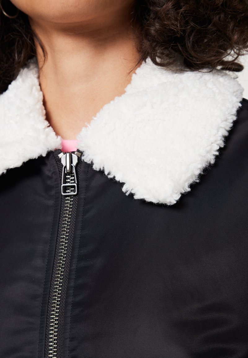 Giubbotto nero con un collo bianco soffice, dotato di una cerniera frontale e una texture elegante. La texture del collo ricorda il morbido pile.