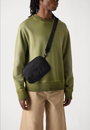 Schwarze Umhängetasche mit strukturierter Oberfläche, rechteckiger Form, einem Tragegurt und einem kleinen geprägten Logo. Getragen über einem olivfarbenen Sweatshirt.