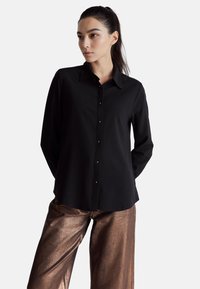 Czarna koszula z długim rękawem, z kołnierzykiem, zapinana z przodu na guziki, o gładkiej fakturze, zestawiona z błyszczącymi, brązowymi szerokimi spodniami.