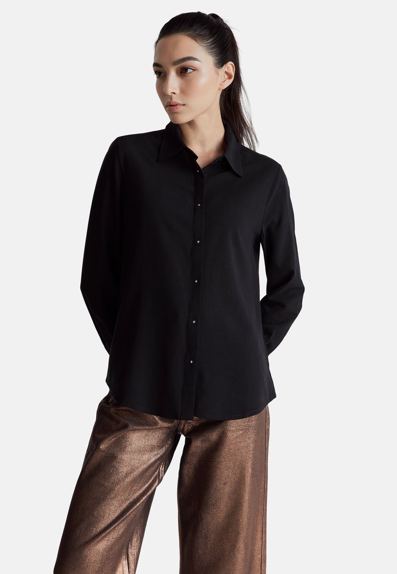 Czarna koszula z długim rękawem, z kołnierzykiem, zapinana z przodu na guziki, o gładkiej fakturze, zestawiona z błyszczącymi, brązowymi szerokimi spodniami.