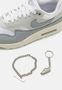 Szürke Nike Air Max cipő fehér fűzőkkel, texturált bőr és hálós felsőrésszel. Ezüst színű lánc karkötő és tornacipő kulcstartó társaságában.