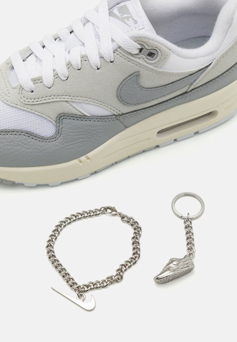 Grijze Nike Air Max schoen met witte veters, gestructureerd leren en mesh bovenwerk. Vergezeld van een zilveren kettingarmband en een sleutelhanger voor sneakers.