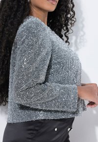 Vrouw met lange gekrulde haren, gekleed in een getextureerde zilveren pailletten top en zwarte satijnen broek met decoratieve stenen, staand in profile.