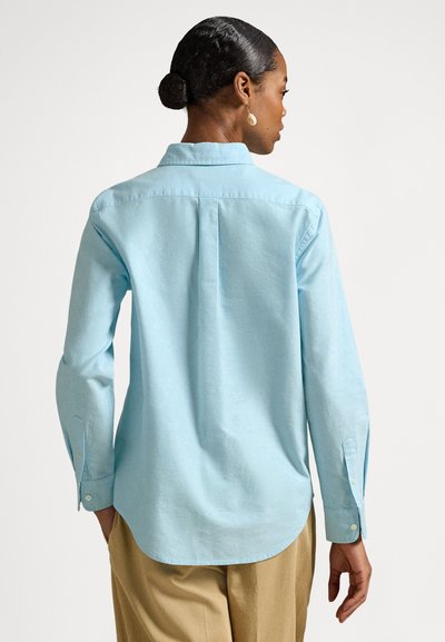 Gaiši zila krekla ar garām piedurknēm, liektu apakšpusi, pogām uz aprocēm un aizmugurējo yoke detaļu. Izgatavots no gludas, viegli teksturētas auduma.