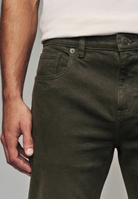 Donkergroene katoenen broek met een gladde textuur, voorzien van een voorzak, knoopsluiting en gestikte details langs de naden.