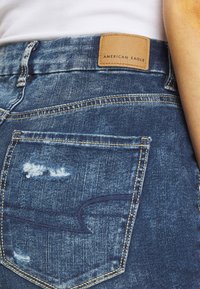 Levi's jeans de mezclilla azul con un diseño desgastado, parche de cuero marrón con "AMERICAN EAGLE" y costuras amarillas en los bolsillos.