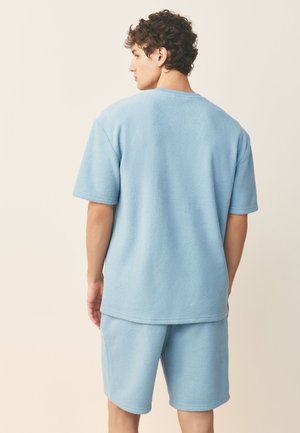 Vyriškas modelis su garbanotais plaukais, vilkintis šviesiai mėlyną trumpomis rankovėmis dėvimą šiltą megztinį ir atitinkamus šortus, stovintis nugara į smėlio spalvos foną.