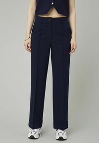 Pantalons large bleu marine avec une texture lisse, des plis pressés, une fermeture par bouton et des ourlets revers, associés à des baskets grises.