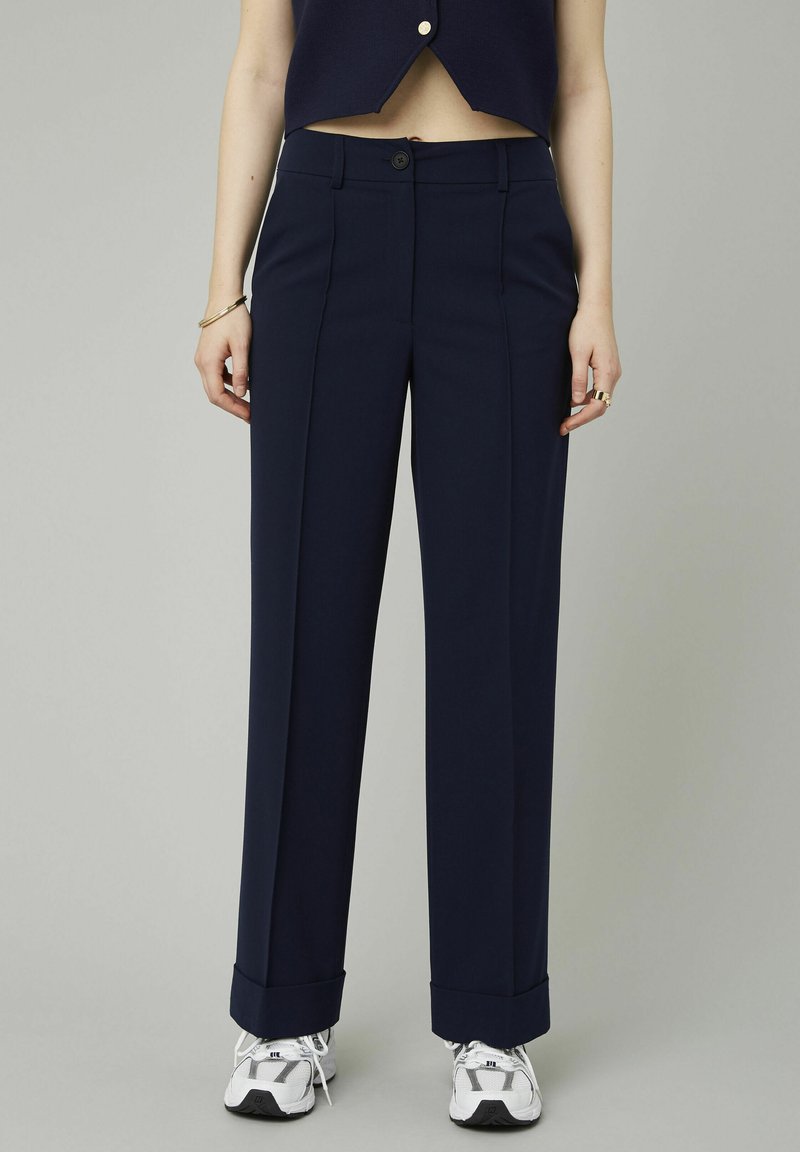 Pantalons large bleu marine avec une texture lisse, des plis pressés, une fermeture par bouton et des ourlets revers, associés à des baskets grises.