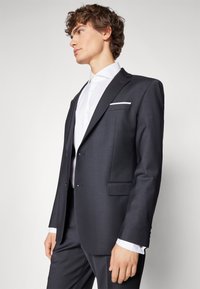 Dark tailored suit with notch lapels, two front pockets, and contrasting white pocket square. Shirt has a classic collar and button front.