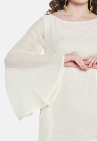 Bijela, lagana, mrežasta majica s zvonastim rukavima. Ima ležeran kroj i teksturiranu tkaninu, s širom izrezom i suptilnim akcentima šivanja.