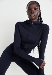 Navy Sportanzug mit eng anliegendem Design, langen Ärmeln und einer mit Reißverschluss versehenen Kapuze. Mit geripptem Textur und Daumenlöchern für zusätzlichen Komfort.