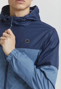 Giacca leggera blu con cappuccio, caratterizzata da diverse sfumature di blu navy e azzurro, polsini elasticizzati e un logo tondo sul petto.