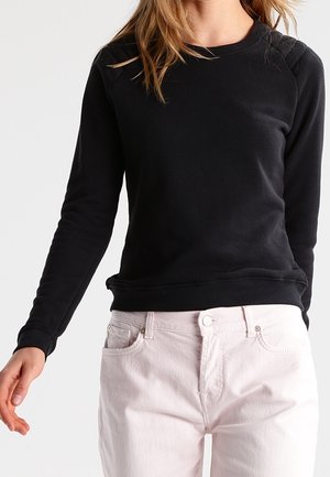 Kvinde iført en ensfarvet sort langærmet skjorte og lyserøde bukser, stående mod en hvid baggrund.