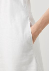 Vestido blanco de lino con un bolsillo lateral; presenta una tela suave y texturizada, y un corte holgado que enfatiza la simplicidad en el diseño.