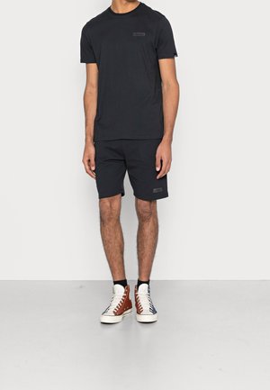 Mężczyzna ubrany w czarną koszulkę z krótkim rękawem i szorty, w kolorowe sneakersy za kostkę, stojący na białym tle.