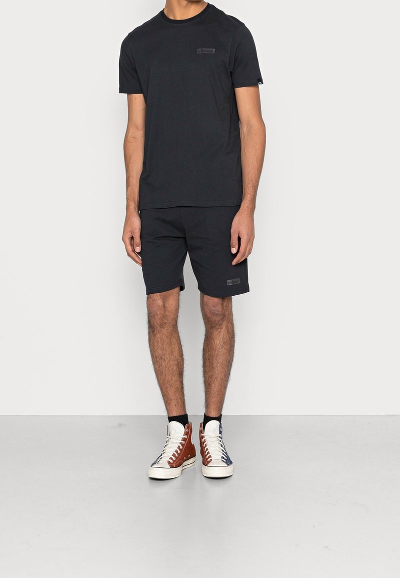 Homme portant un t-shirt à manches courtes noir et un short, avec des baskets montantes à blocs de couleurs, debout devant un fond blanc uni.