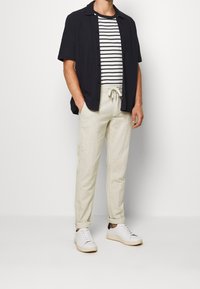 Marinblå kortärmad skjorta med knapp över en svartvit randig t-shirt och ljusbeige byxor, i kombination med vita sneakers.