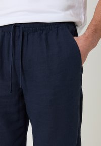 Hombre con pantalones azul marino con cordón y la mano en el bolsillo derecho, combinado con una camisa blanca metida de manera suelta en la cintura.