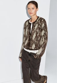 Cardigan lavorato a maglia in marrone e crema con un motivo testurizzato, scollatura rotonda e chiusure con bottoni; abbinato a pantaloni larghi scuri.