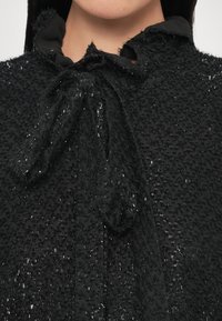 Blouse noire texturée avec un col à volants, dotée d'une fermeture à nouer. Le tissu comporte des accents brillants subtils et une apparence douce et superposée.