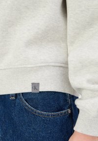 Světlé šedé triko s žebrovaným lemem, na kterém je malá šedá visačka s logem "cK", nošené přes modré džínové kalhoty s detaily kapes.