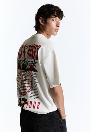 Jeune homme aux cheveux foncés portant un t-shirt blanc de la tournée de Guns N' Roses et un pantalon noir, regardant par-dessus son épaule sur fond blanc.