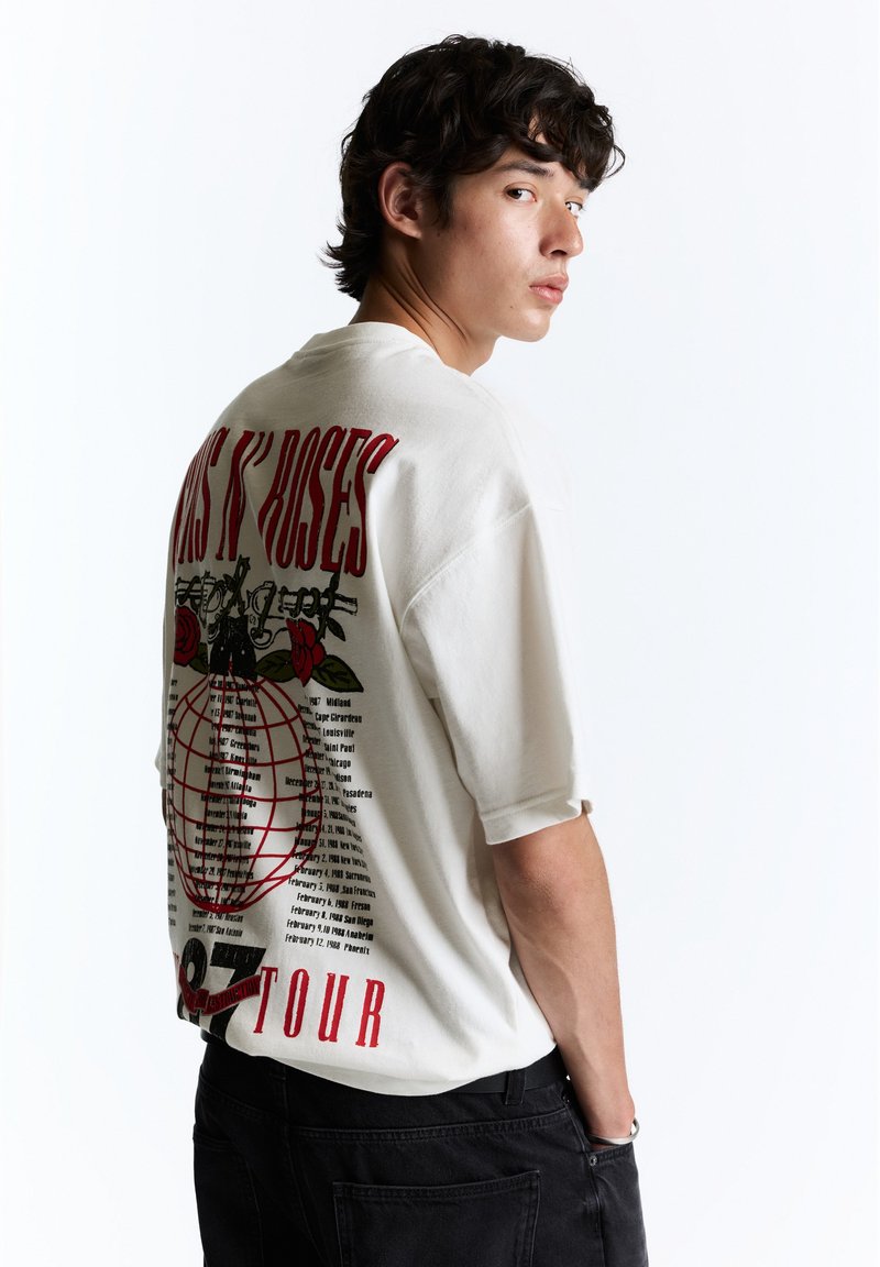 Jeune homme aux cheveux foncés portant un t-shirt blanc de la tournée de Guns N' Roses et un pantalon noir, regardant par-dessus son épaule sur fond blanc.