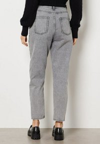 Henkilö, joka on pukeutunut korkeavyötäröisiin harmaisiin farkkuihin, joissa on takataskut, mustiin paksupohjaisiin loafereihin ja mustaan pitkähihaiseen neuleeseen, seisoo sisätiloissa.