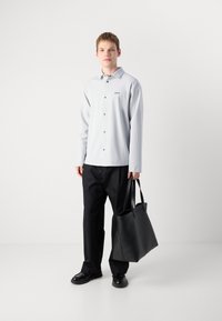Lys grå langermet skjorte med knappefront og svart logo, kombinert med svarte bukser og en matt svart tote bag med håndtak.