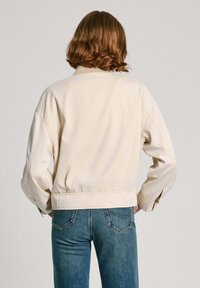 Jasna kremowa krótka kurtka o luźnym kroju, z szerokimi ramionami i zapiętymi mankietami, w połączeniu z niebieskimi dżinsami.
