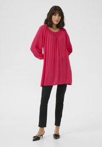 Blouse longue plissée rose avec un col rond et des détails froncés, associée à un pantalon noir slim et des mules à bout pointu ornées de nœuds.