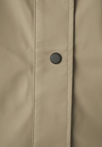 Tejido de impermeable beige con una textura suave. Presenta un botón de presión negro y costuras en contraste a lo largo del pliegue delantero.