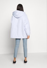 Giacca oversize azzurro chiaro con cappuccio, polsini elastici e una texture liscia, abbinata a denim chiaro e sneakers nere.