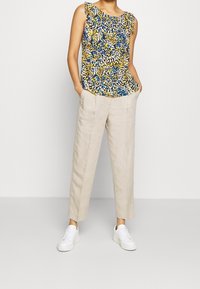 Blouse sans manches à motifs bleus, jaunes et blancs associée à un pantalon en lin beige et des baskets blanches ; caractérisée par une coupe décontractée et des poches latérales.