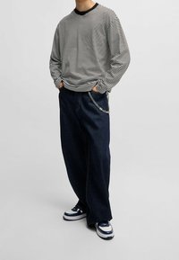 Person in schwarz-weiß gestreiftem Langarmshirt, lockeren dunkelblauen Jeans mit Kette und weißen sowie marineblauen Sneakern vor einem schlichten Hintergrund stehend.