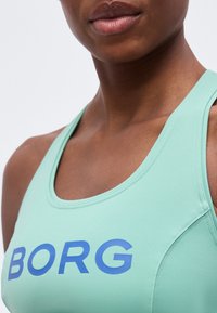 Κοντινό πλάνο ενός ατόμου που φοράει ανοιχτοπράσινο αθλητικό τοπ με μπλε κείμενο "BORG" στο στήθος, δείχνοντας λαιμό και ώμους.