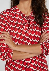 Blouse à motifs rouges avec un design géométrique, des accents blancs et rouge foncé, un col à nouer, des détails en boutons et des manches retroussées.