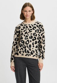 Pull beige avec un motif léopard noir. Présente un col rond, des poignets à côtes et une coupe décontractée, associé à un pantalon gris foncé.