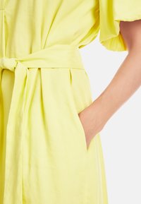 Helles gelbes Kleid mit gebundenem Taillenzug, weichem Stoff, dezenten Taschen und weiten Ärmeln mit sanften Falten für zusätzliche Bewegungsfreiheit.