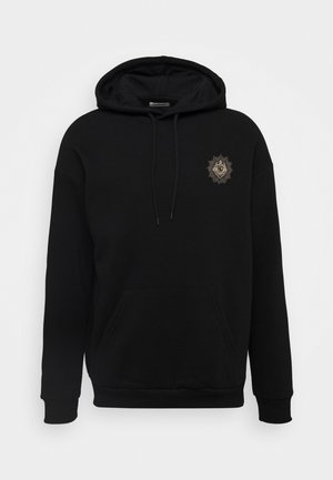 Homme portant une veste en peau retournée marron par-dessus un sweat à capuche noir, un pantalon beige aux revers retroussés et des baskets montantes mauves avec des lacets et des semelles blancs.