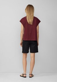 Burgunderfarbendes T-Shirt mit kurzen Ärmeln, kombiniert mit hochgekrempelten schwarzen Jeansshorts. Das Outfit wird durch flache schwarze Sandalen ergänzt. Schlichtes Design, lässige Passform.