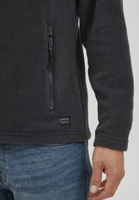 Fleece-pullover i mørkegrå med glidelås-lomme, strukturert stoff og merket etikett. Har en avslappet passform og minimalistisk design.