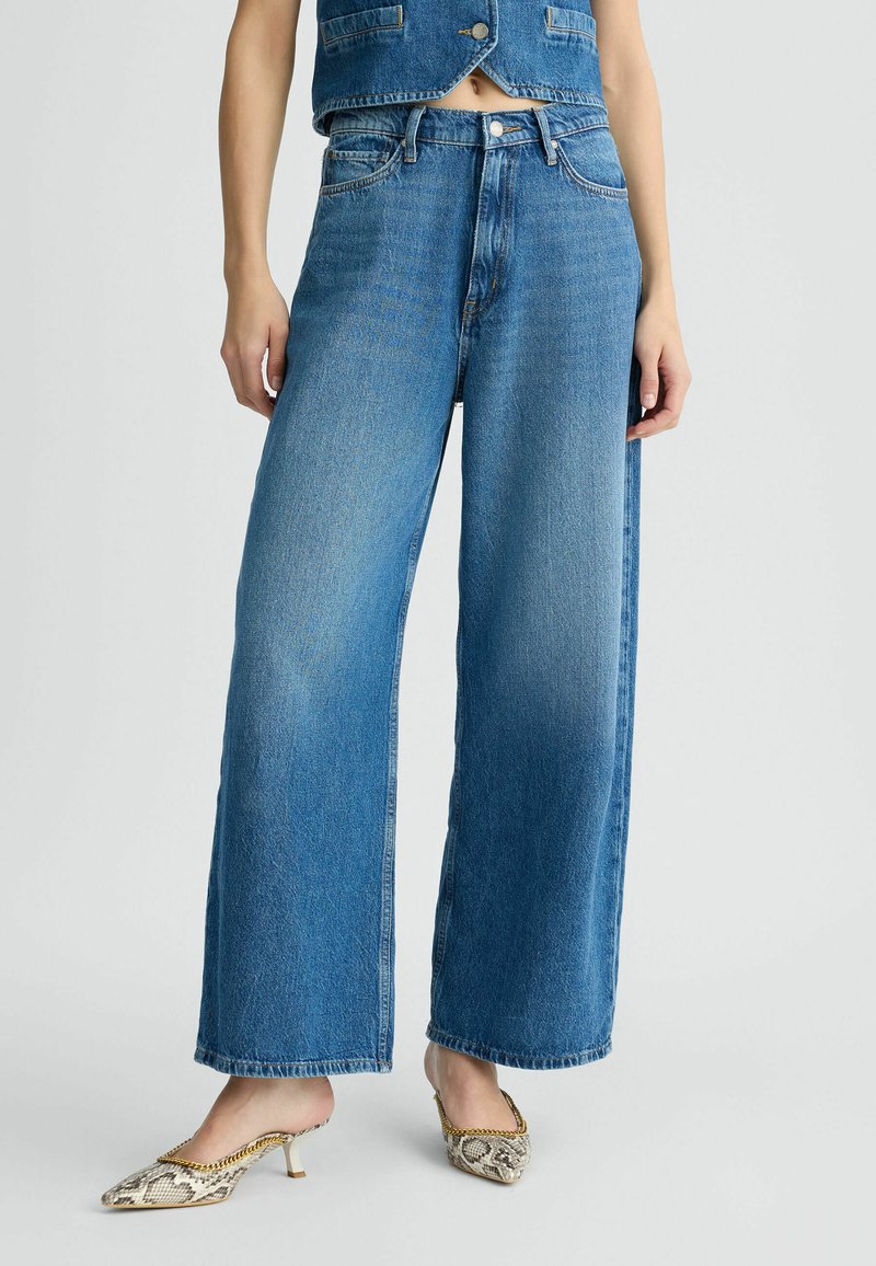 Laia denim püksid, mis on puusalt laiade jalgadega ja kõrge vöökohaga, kergelt pleekinud ja toorena servadega, koos kraapi mustriga mules'idega, millel on väike konts.