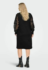 Sort kjole med lange, gjennomsiktige volangermet, ledig passform og knelang design. Har teksturert stoff og enkel halslinning.