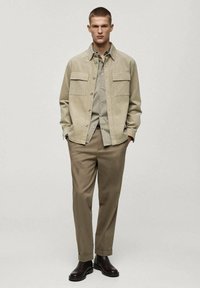 Giacca camicia in corduroy beige chiaro con due tasche sul petto, indossata su una camicia grigia chiaro, abbinata a pantaloni beige e stivali marrone scuro.