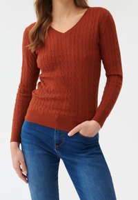 Femme portant un pull en maille torsadée couleur rouille à col en V et un jean bleu, une main dans la poche, debout devant un fond blanc.